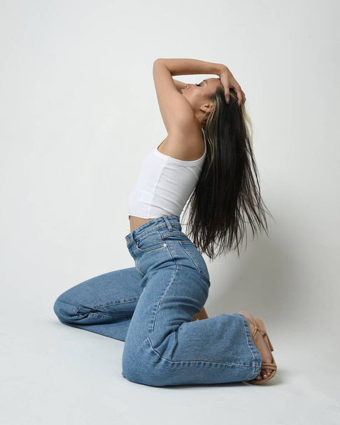 Full length portrait of brunette female asian model wearing casual clothes, white singlet shirt, denim jean pants. Comfortable seated pose. Isolated on studio background. - Photo, Image