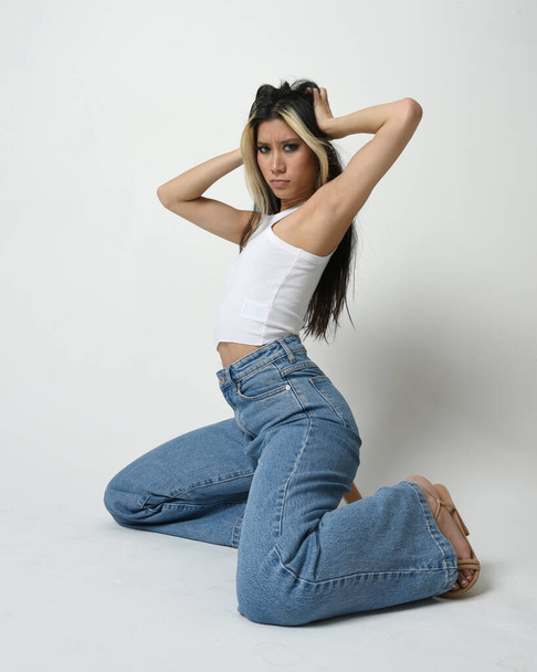 Портрет брюнетки жіночої азіатської моделі в повсякденному одязі, біла сорочка-синглет, джинсові штани. Зручна сидяча поза. Ізольовані на студійному фоні. - Фото, зображення