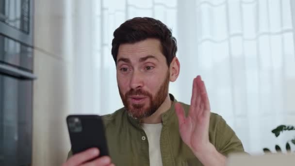 Homme brune caucasien avec barbe tenant smartphone et disant bonjour sur appel vidéo tout en agitant la main. Homme souriant utilisant gadget moderne et Internet pour la communication en ligne et le travail de la maison. - Séquence, vidéo