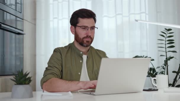 Portrét pohledného vousatého muže sedícího v moderním obývacím pokoji s laptopem, jak otáčí hlavu a usmívá se do kamery. Šťastný muž na sobě ležérní oblečení a brýle těší práci na pohodlném domácím pracovišti. - Záběry, video