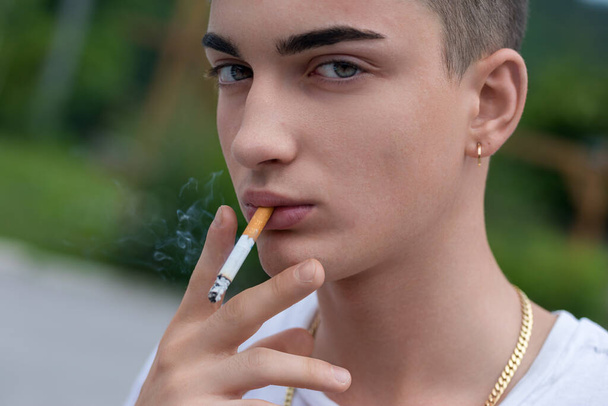 Defiant arcú fiatal felnőtt élvezi a cigaretta, ékszerek díszítik a fül és a nyak, közepette elmosódott háttér - Fotó, kép