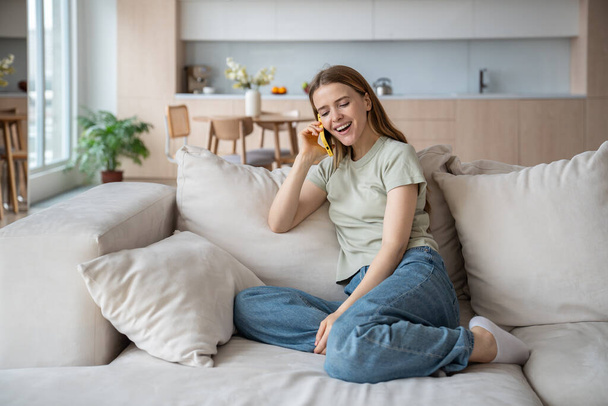 Boldog nő élvezze a kellemes beszélgetést okostelefonon pihentető ül a kanapén otthon. Egy lány telefonál, mosolyog, vicces sztorikat hallgat a barátjától. Mobilkommunikáció, jó hírek. - Fotó, kép