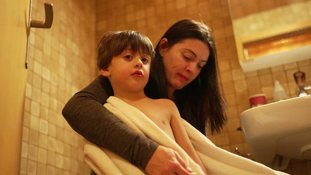 Mother kissing son on the cheek and drying body wrapped in towel after bath time. Authentic parent and child loving affection and caring relationship - Photo, Image