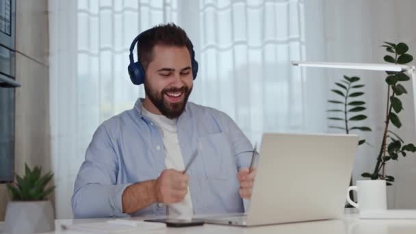 Playful freelancer in wireless headset dancing and pretending drumming with pencils in front of desktop with pc. Cheerful man making break from work routine and fooling around with favourite music. - Footage, Video