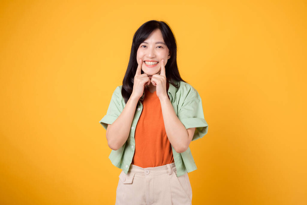 Asiatico allegra donna 30s indossa camicia verde e arancione. Con le dita rivolte ai denti, sottolinea la salute dentale e il sorriso radioso isolato su sfondo giallo. promuovere il concetto di cura orale. - Foto, immagini