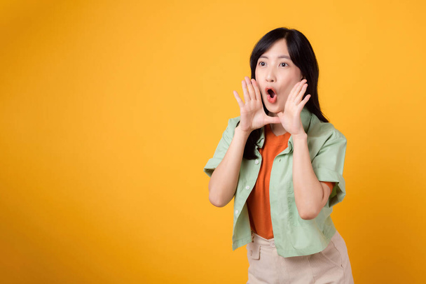 Femme asiatique joyeuse trentaine portant une chemise orange et une chemise verte montrant un geste de main couvrant la bouche, symbolisant un cri ou une surprise. Moments énergétiques et concept d'exubérance. - Photo, image