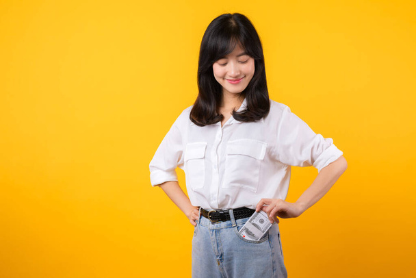Imagem cativante da jovem mulher asiática vestindo camisa branca e jeans jeans jeans, segurando com confiança dinheiro de seu bolso da camisa. Moda atende empoderamento financeiro. Perfeito para temas financeiros e de moda. - Foto, Imagem