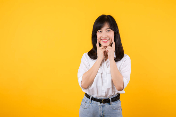 Mladá asijská krásná žena šťastný úsměv na sobě bílou košili a džínové rostliny přes žluté izolované pozadí s úsměvem sebevědomě ukazuje a ukazuje prsty zuby a ústa. Zubní koncept. - Fotografie, Obrázek