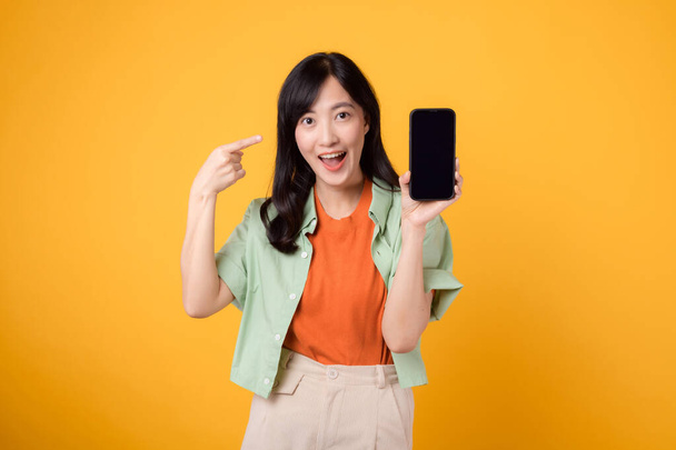 Vrolijke Aziatische vrouw in de 30 met een groen en oranje shirt. wijst vinger naar smartphone, symbool voor connectiviteit en digitale empowerment. technologie en moderne levensstijl op een levendige gele achtergrond. - Foto, afbeelding