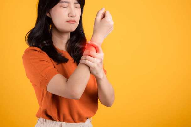 Eine Frau in den 30er Jahren trägt ein orangefarbenes Hemd, während sie ihr Handgelenk vor Schmerzen hält. Veranschaulichung der Herausforderungen der Handgelenksarthritis, dient als Konzept für Gesundheit und Wellness. - Foto, Bild