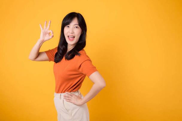 Promieniująca pozytywność, azjatycka wesoła kobieta po trzydziestce w pomarańczowej koszuli pokazuje w porządku gest dłoni na żółtym tle, symbolizujący aprobatę i pewność. Pozytywność, porozumienie, koncepcja zaufania. - Zdjęcie, obraz