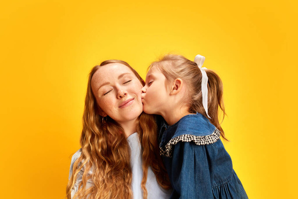 Little girl, daughter cissing on cheek her beautiful happy mother against yellow studio background. Bonding moment. Concept of Mothers Day, International Happiness Day, family, love and care - Foto, Imagem