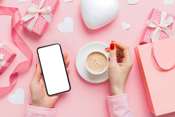 Female hands holding smart phone with coffee for Valentine day, gift box and envelope, hearts. - Foto, Bild