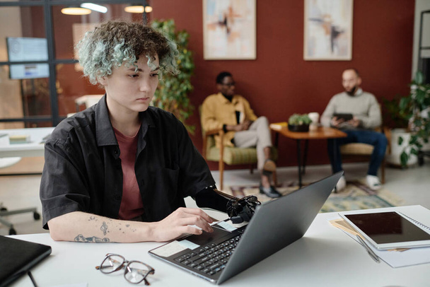 Young girl with prosthetic arm browsing laptop at office, her male colleagues sitting on background - Photo, Image