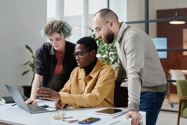 Молодые офисные работники изучают информацию о ноутбуке, черный разговаривает со своими коллегами - Фото, изображение