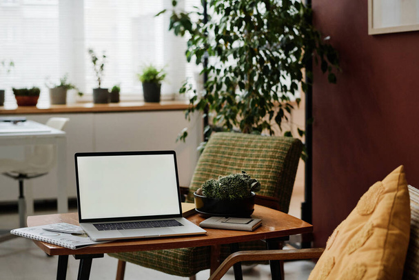 Laptop with white screen mockup placed on table in modern office with greenery and vintage furniture - Photo, Image