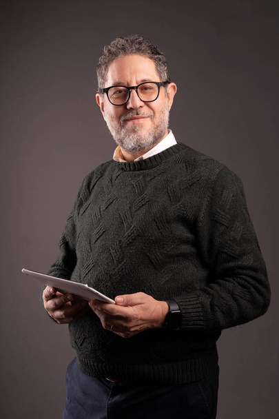 Homme d'affaires mature en tenue décontractée utilisant avec confiance une tablette numérique, debout sur un fond gris. - Photo, image