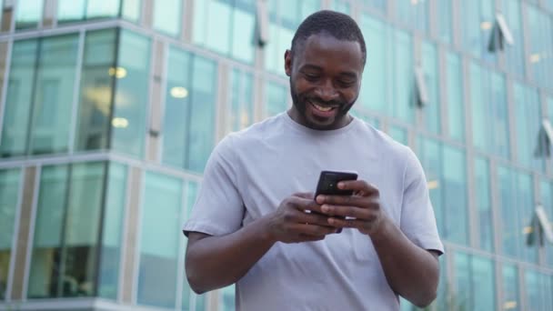 Ευτυχισμένος Αφροαμερικανός άνδρας ευφορική νικητής με smartphone στο δρόμο στην πόλη. Άνθρωπος πρόσωπο κοιτάζοντας το κινητό τηλέφωνο ανάγνωση μεγάλη είδηση πάρει καλό αποτέλεσμα κερδίζοντας σε απευθείας σύνδεση αίσθημα προσφοράς κατάπληκτος. Κερδίζοντας χειρονομία - Πλάνα, βίντεο