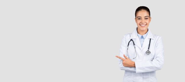 Eine strahlende europäische Ärztin mit einem zur Seite zeigenden Stethoskop, das auf ein medizinisches Produkt oder eine Dienstleistung hindeutet, mit freundlichem Auftreten auf hellgrauem Hintergrund, Panorama - Foto, Bild