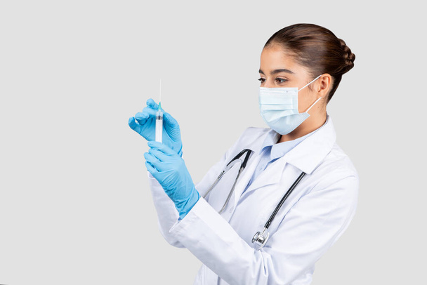 Médico europeu concentrado em jaleco e máscara protetora, examinando cuidadosamente uma seringa, ilustrando a precisão médica e os serviços de vacinação, isolado em fundo de estúdio cinza - Foto, Imagem