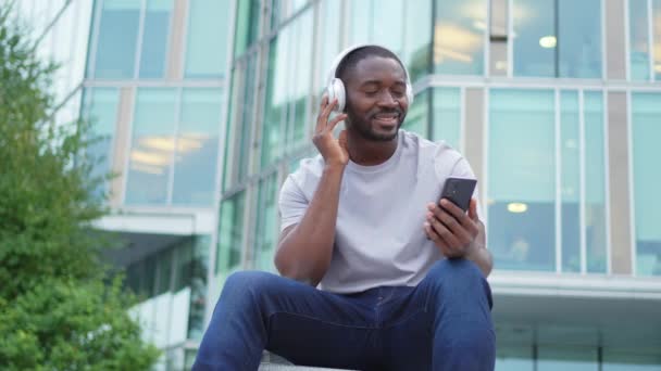 Music song concept. Good start day. Happy African American man listening modern hit on street in city. Person guy wearing headphones enjoy listening favorite music on phone. People lifestyle joy - Footage, Video