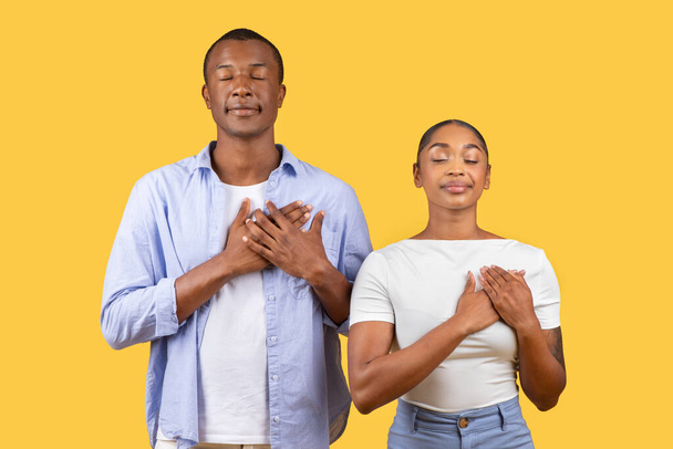 Friedliches schwarzes Paar steht mit Händen über dem Herzen und geschlossenen Augen in Geste der Dankbarkeit und Besinnung vor beruhigendem gelben Hintergrund - Foto, Bild