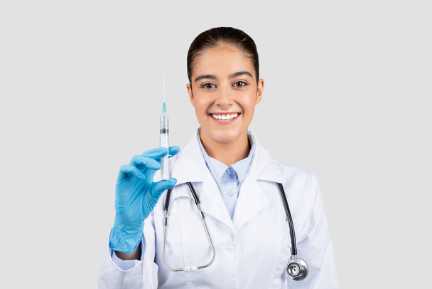 Beyaz önlüklü ve mavi eldivenli neşeli Avrupalı bir bayan doktor, gri stüdyo arka planında izole edilmiş tıbbi tedaviler ve aşılar konusunda uzmanlığını temsil eden bir şırınga sergiliyor. - Fotoğraf, Görsel