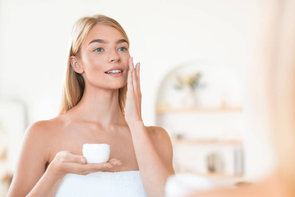 Aantrekkelijke blonde dame geniet van huidverzorging terwijl brengt moisturizer op haar gezicht thuis badkamer, verzorging van de huid en verwennerij haar schoonheid in de ochtend, staande verpakt in handdoek - Foto, afbeelding