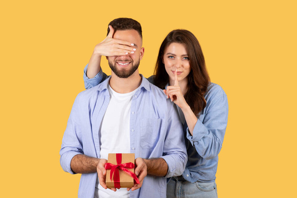 Γυναίκα καλύπτει τα μάτια των ανδρών για δώρο έκπληξη στιγμή, το δάχτυλό της στα χείλη της για μυστικότητα, καθώς κρατά δώρο με κόκκινη κορδέλα, σε κίτρινο φόντο - Φωτογραφία, εικόνα