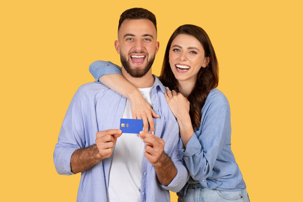 Feliz joven pareja compartiendo risa mientras mantiene unida la tarjeta de crédito, lo que sugiere una divertida experiencia de compra, frente a un cálido fondo amarillo - Foto, imagen