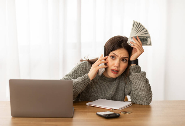 Aile bütçesi üzerinde çalışan sıkıntılı genç kadın ev hanımı, dizüstü bilgisayarın önünde oturuyor, elinde para tutuyor, telefonla konuşuyor ve kocasına dokunuyor. İflas, kriz - Fotoğraf, Görsel