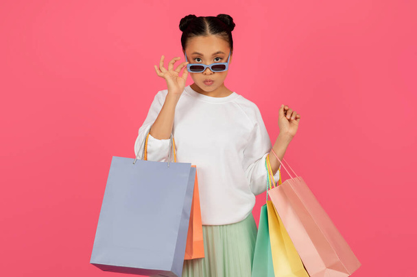 Alışveriş torbalı şık Asyalı kadın güneş gözlüğünü ayarlıyor ve kameraya bakıyor, alışveriş yapan genç Koreli bayan, moda ve satışlardan keyif alıyor, pembe arka planda tek başına duruyor. - Fotoğraf, Görsel
