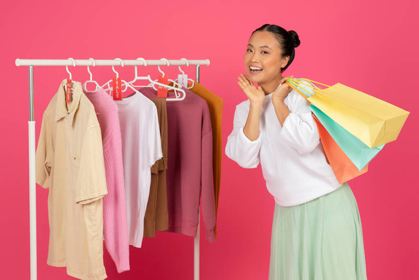 Concept de vente. Femme asiatique excitée tenant des sacs à provisions colorés tout en se tenant près du porte-vêtements, joyeuse coréenne heureuse de nouveaux vêtements, posant sur fond de studio rose, gros plan - Photo, image