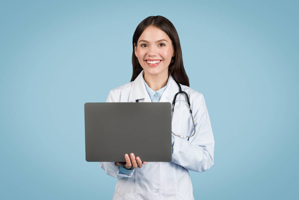 Jovem médica alegre com estetoscópio, segurando computador portátil como símbolo dos serviços de telessaúde e gerenciamento digital de pacientes - Foto, Imagem
