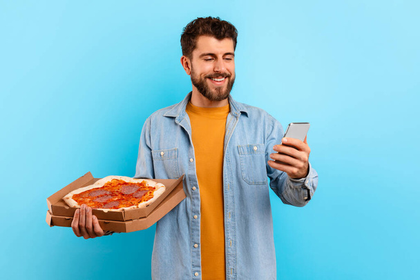 Joven hambriento sosteniendo la caja de pizza y usando un teléfono inteligente parado sobre el fondo azul del estudio, ordenando la entrega de comida chatarra del restaurante pizzería en la aplicación móvil - Foto, imagen