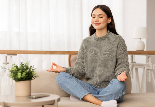 Entspannte attraktive junge Frau in Lotus-Pose mit geschlossenen Augen auf dem heimischen Sofa sitzend, meditierend, Händchenhaltend in Zen-Geste, Ruhe genießend, Raum kopierend. Entspannung, Stressabbau - Foto, Bild