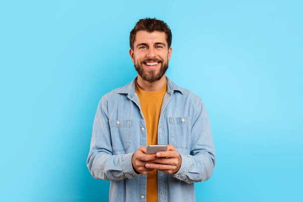 Iloinen kaveri älypuhelimen katselee kameraa ja hymyilee, tekstailee sosiaalisessa mediassa sinisen studion taustalla. Positiivinen nuori mies rento chattailuun puhelimessa, käyttämällä uutta mobiilisovellusta - Valokuva, kuva