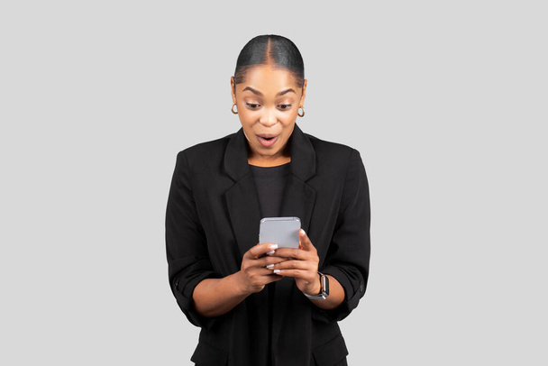 Sorpresa e deliziata giovane donna d'affari legge un messaggio sul suo smartphone, la sua espressione indica buone notizie o un'interazione positiva, isolata su sfondo grigio studio - Foto, immagini