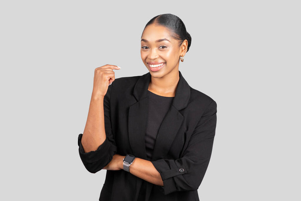 Χαρούμενη νεαρή Αφροαμερικάνα με μαύρο σακάκι, έξυπνη στον καρπό, κάνει μια δυνατή χειρονομία με χαμόγελο, συμβολίζοντας την αυτοπεποίθηση και την επιτυχία, απομονωμένη σε γκρι φόντο στούντιο - Φωτογραφία, εικόνα