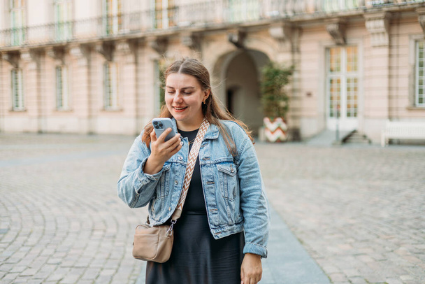 Позитивная весёлая женщина-туристка, одетая в повседневную одежду, держит смартфон-гаджет для общения на городской улице. Женщина болтает на мобильном смартфоне во время отдыха на открытом воздухе, концепция городского образа жизни - Фото, изображение