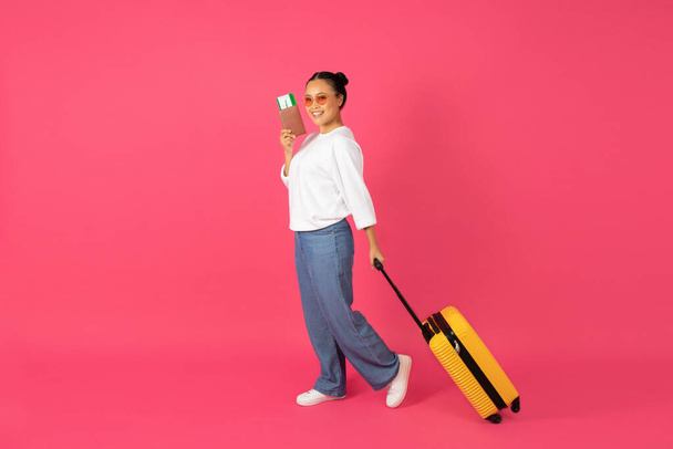 Onnellinen aasialainen matkustaja nainen yllään aurinkolasit kävely passi ja matkalaukku pinkl taustalla, hymyilevä korealainen nainen vetää matkatavaroiden laukku, symboloi tyylikäs ja valmis matka, kopioi tilaa - Valokuva, kuva