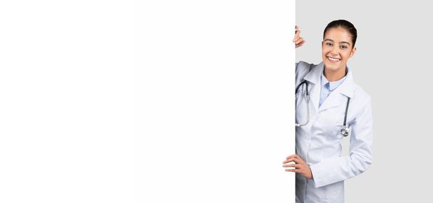 Счастливая кавказская женщина-врач тысячелетия в белом халате с огромным баннером с свободным пространством, изолированная на сером фоне студии, панорама. Медицинская презентация, здравоохранение, продажа, внимание и реклама - Фото, изображение