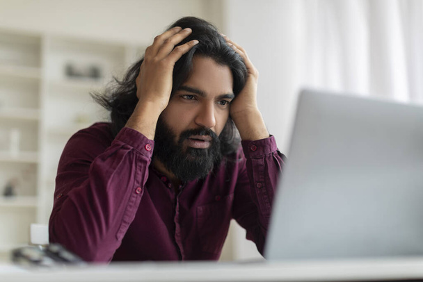 Hombre indio preocupado agarrando la cabeza en la frustración mientras mira fijamente a la pantalla de la computadora, estresado joven hombre oriental que sufre problemas de trabajo, sentado en el escritorio en la oficina en casa, disparo de primer plano - Foto, imagen