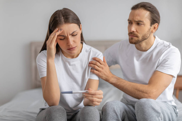 Smutny nieszczęśliwy mąż uspokaja żonę patrząc na test ciążowy, siedząc w łóżku. Niepłodność, niechciana ciąża, problemy zdrowotne, planowanie rodziny i wsparcie - Zdjęcie, obraz