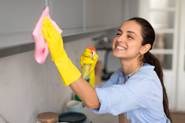 黄色い手袋とカジュアルな服の放射状の若い女性は,ピンクの布でキッチンの表面をエネルギー的に磨き,満足している - 写真・画像