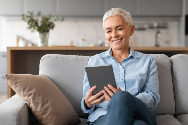Usmívající se běloška zralá žena s digitálním tabletem, sedící s počítačovými hračkami a odpočívající na pohovce v moderním domácím interiéru o víkendu. Lady prohlížení webových stránek a internet uvnitř - Fotografie, Obrázek