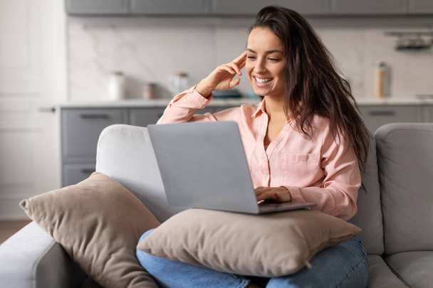 Mujer joven relajada y alegre con una sonrisa encantadora que se conecta con su computadora portátil mientras se sienta con las piernas cruzadas en un acogedor sofá en casa, emanando un ambiente de elegancia casual - Foto, imagen