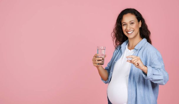 Uśmiechnięta młoda, zdrowa kobieta w ciąży trzymająca pigułkę i szklankę wody, oczekująca, że pani przyjmie codziennie suplementy witamin medycyny dla ochrony zdrowia w czasie ciąży, różowe tło, przestrzeń do kopiowania - Zdjęcie, obraz