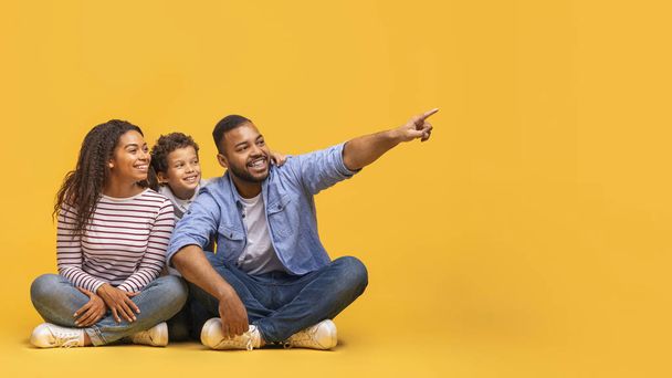 Hieno tarjous. Onnellinen musta vanhemmat ja pieni poika istuu lattialla ja osoittaa viereen Copy Space, iloinen Afrikkalainen amerikkalainen perhe kolme osoittaa vapaa paikka mainos suunnittelu, Panorama - Valokuva, kuva