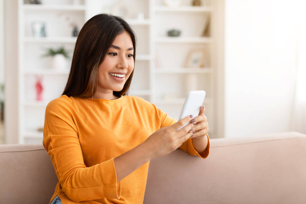 Szczęśliwa atrakcyjna milenijna Chinka relaksująca się na kanapie z telefonem w rękach, przewijanie, websurfing, sprawdzanie miłej oferty online, deal, za pomocą aplikacji mobilnej, home interior, copy space - Zdjęcie, obraz
